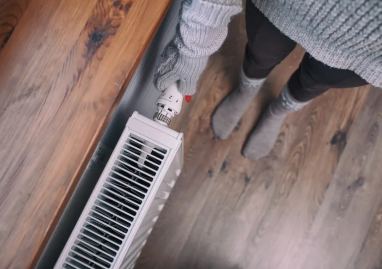 Person wearing gray sweater and gloves adjusting a white radiator valve, standing on a wooden floor.